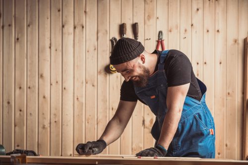 Experienced carpenter in work clothes and small buiness owner working in woodwork workshop,  using sandpaper for polishing  wooden bar at worktable in workshop