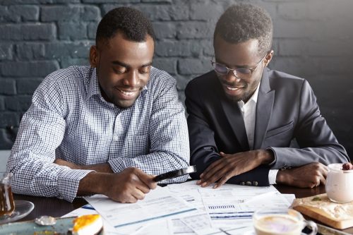 Happy African American office workers dressed in formal clothing having cheerful looks, studying and amalyzing legal documents on table using magnifying glass while getting papers ready for meeting