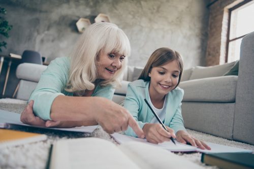 Photo of two people white haired grandma small granddaughter lying comfy floor near couch homework studying writing diary spending weekend together house indoors