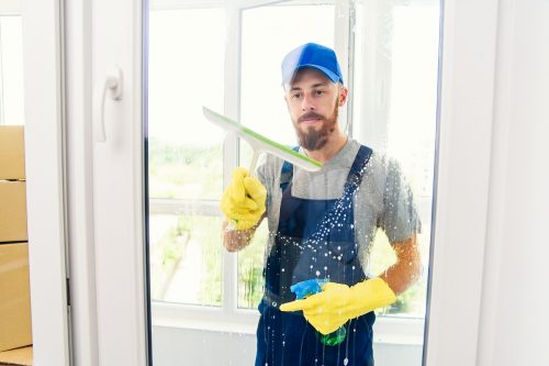 Handsome male janitor from cleaning company using a squeegee to clean a window in an apartment wearing special uniform and rubber gloves.