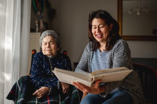 Old sick woman with memory loss sitting in wheelchair. Smiling daughter holding a photo album trying to remember.