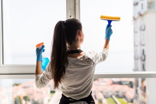 people, housework and housekeeping concept - happy woman in gloves cleaning window with rag and cleanser spray at home